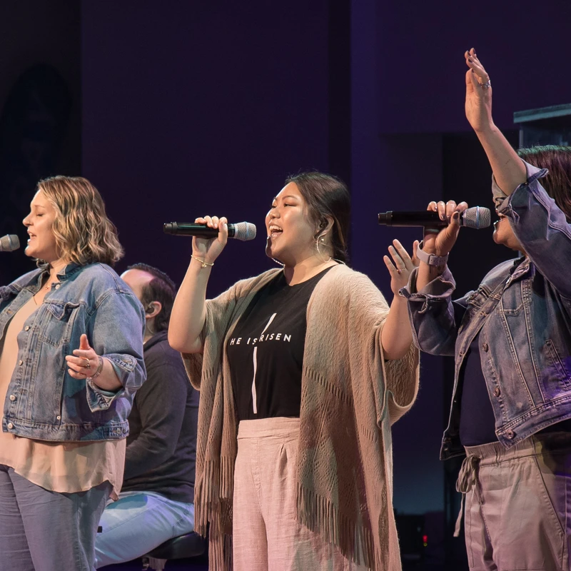 Three women sing into microphones on a dimly lit stage. The center woman wears a 'He Is Risen' shirt, while the woman on the right raises her hand in praise.
